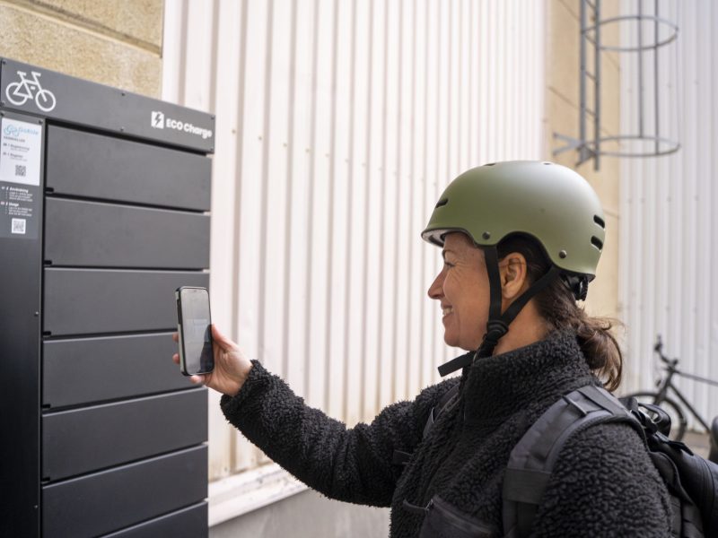 A person wearing a bicycle helmet uses a cell phone at an outdoor ECO Charge to open or reserve a compartment.