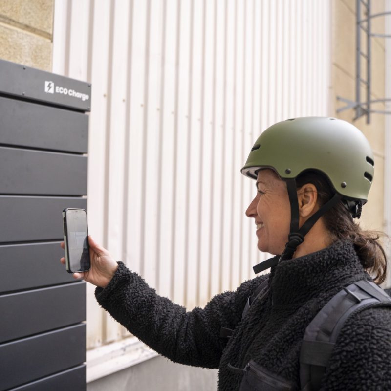 A person wearing a bicycle helmet uses a cell phone at an outdoor ECO Charge to open or reserve a compartment.