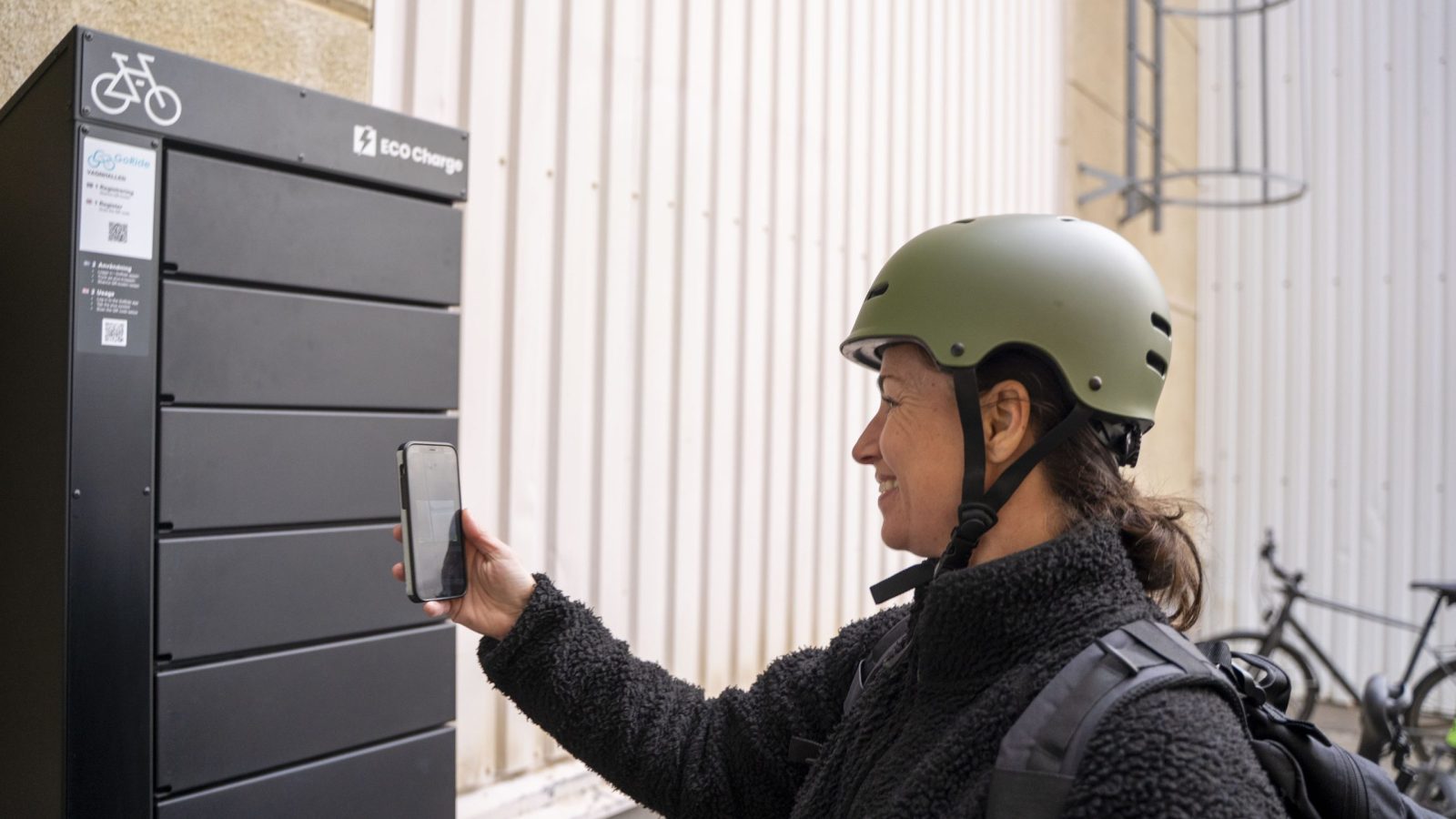A person wearing a bicycle helmet uses a cell phone at an outdoor ECO Charge to open or reserve a compartment.